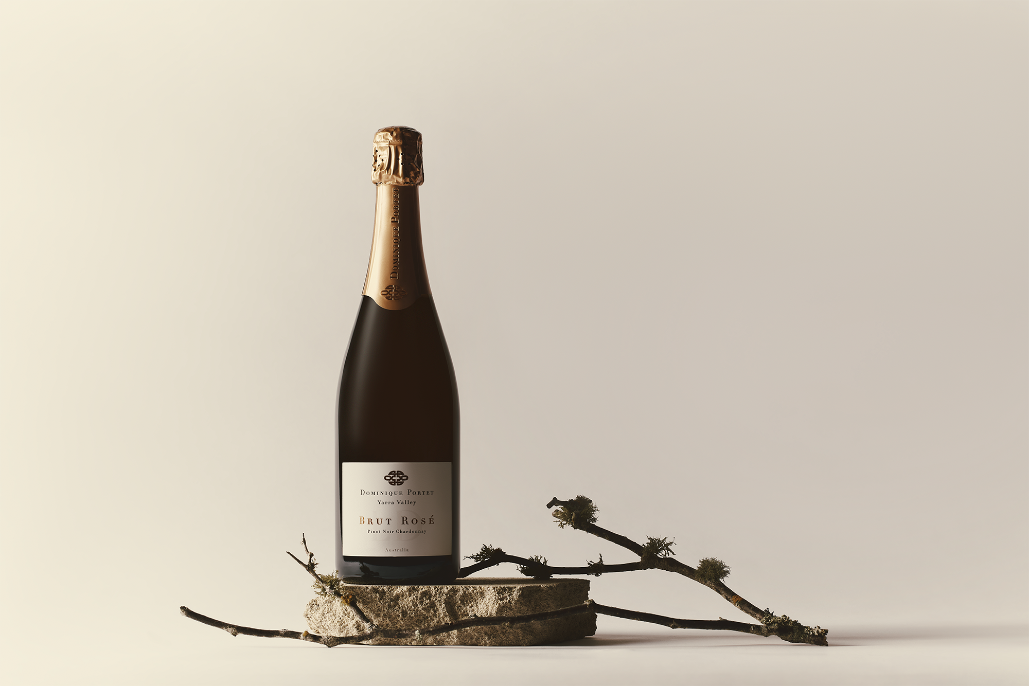 A bottle of sparkling brut rosé wine stands on a rough stone slab, accompanied by a twig with small mossy growths, set against a neutral beige background.