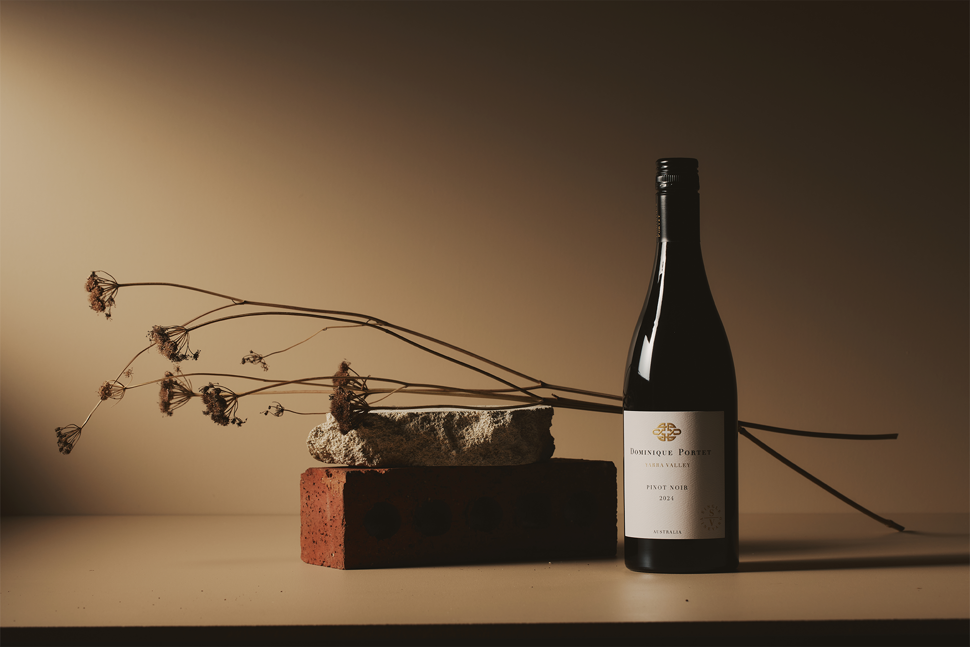 A bottle of wine stands beside a red brick and a gray stone, with dried flowers arranged behind them, set against a beige background with dramatic lighting.