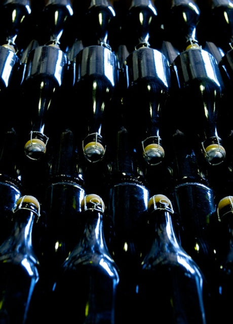 Rows of dark glass bottles are stacked closely together, viewed from below. The bottles have metal clasps and stoppers, reflecting light, creating a pattern of shiny highlights and deep shadows.
