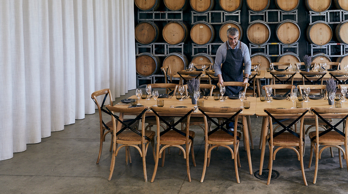 A man in an apron sets a long wooden dining table with glassware and plates in front of stacked wooden barrels in a winery or tasting room.