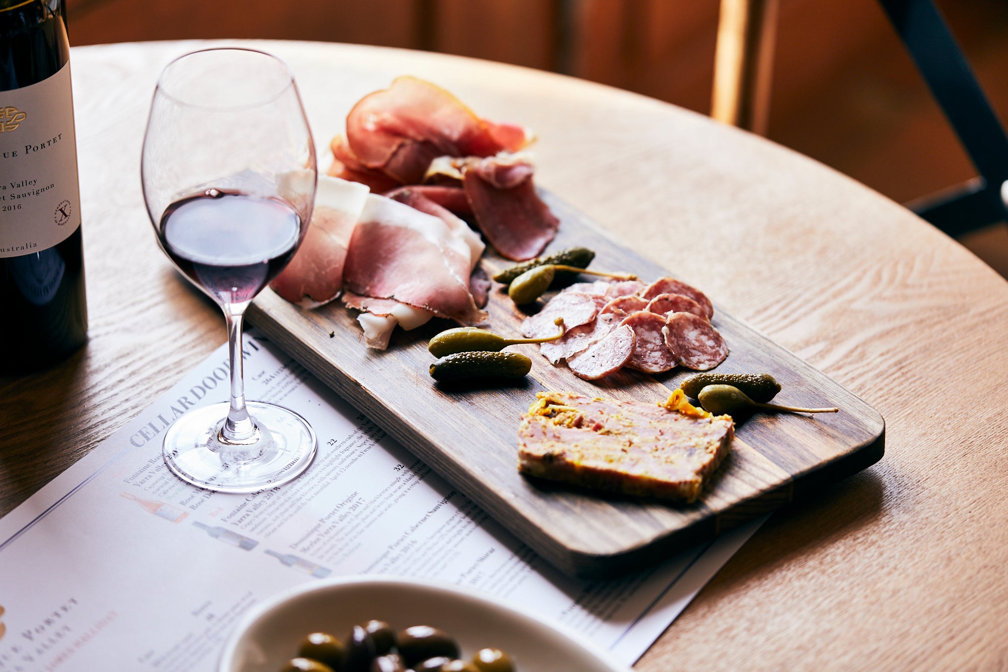 A wooden board with assorted cured meats, pickles, and pâté sits on a table next to a glass of red wine, with a wine bottle and a bowl of olives partially visible nearby.