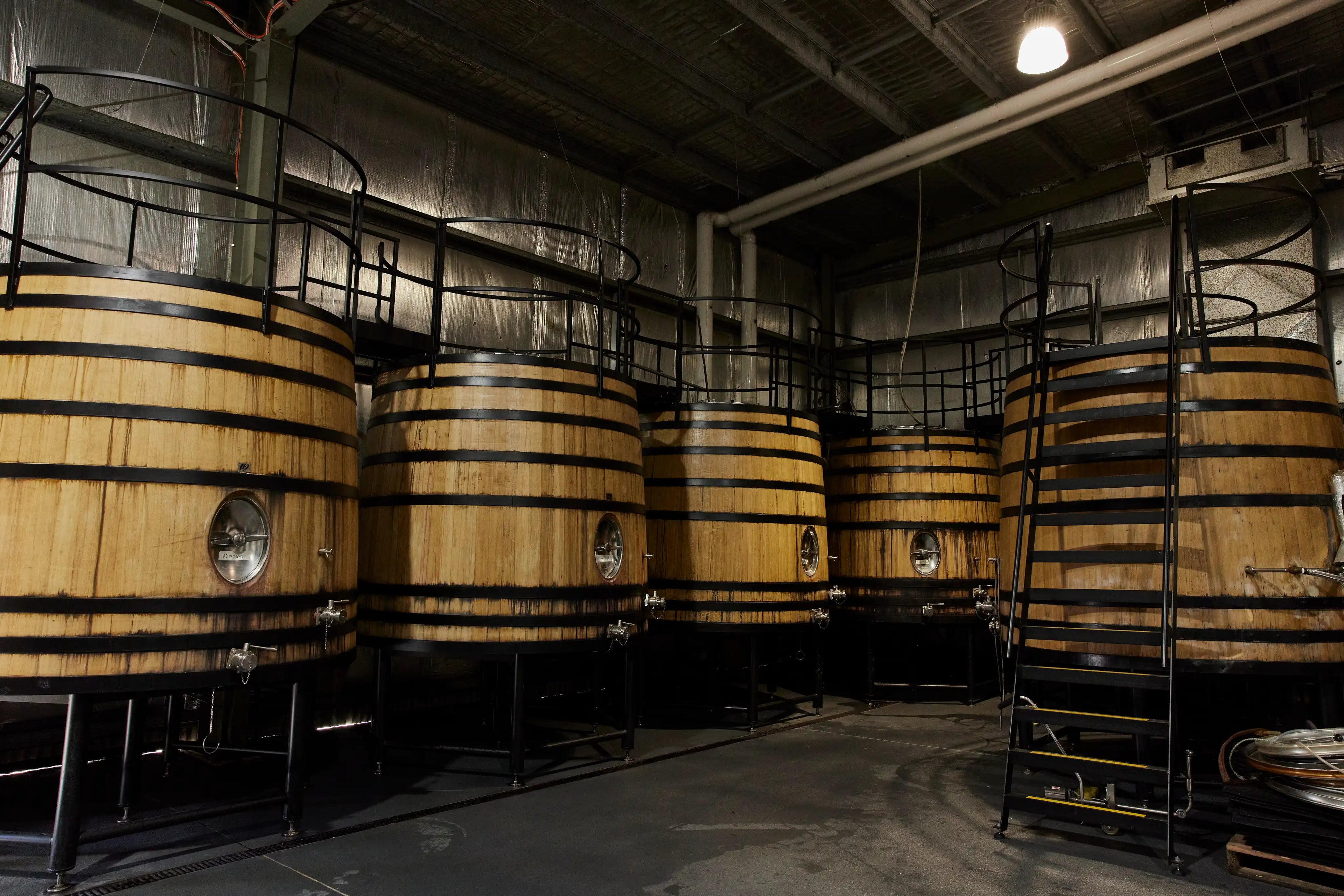 Large wooden fermentation tanks with black metal bands are lined up inside a dimly lit industrial brewery or distillery, with metal railings and walkways above each tank.