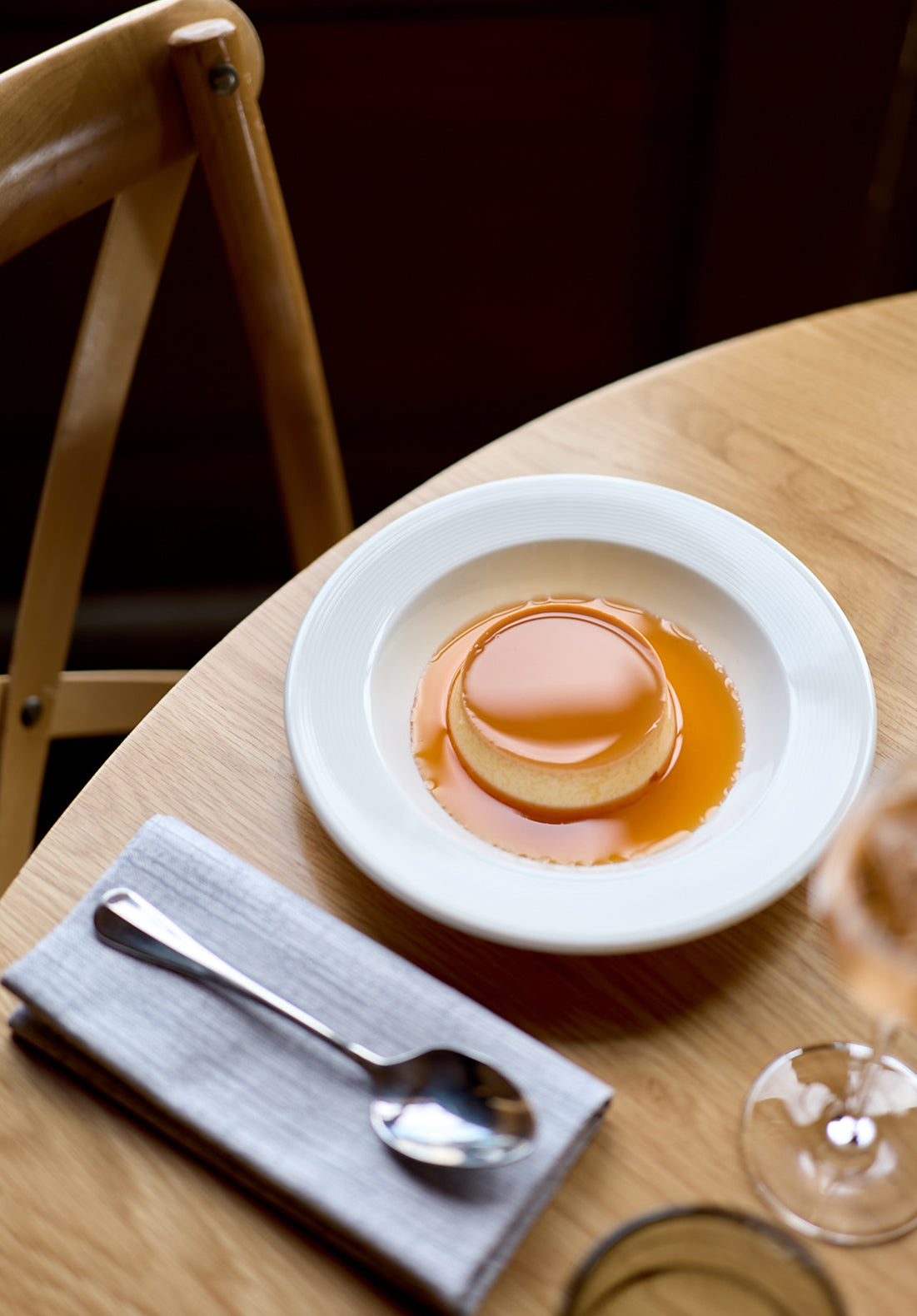 A round caramel flan dessert sits on a white plate atop a wooden table, next to a folded gray napkin and a spoon. A wooden chair and part of a wine glass are also visible.